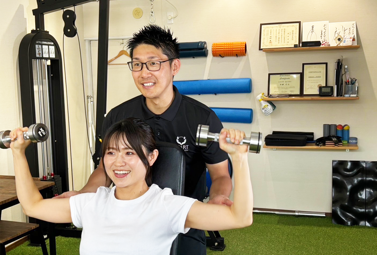 女性がダンベルを使ってトレーニングをし、トレーナーがサポートしている様子